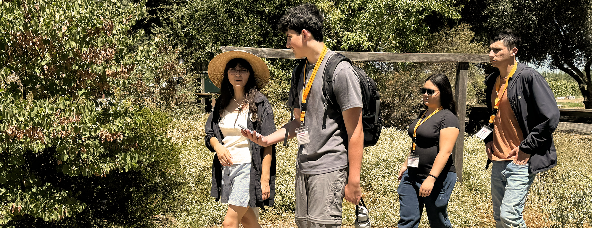 Five people walking a trail through green shrubs, one wearing a straw hat