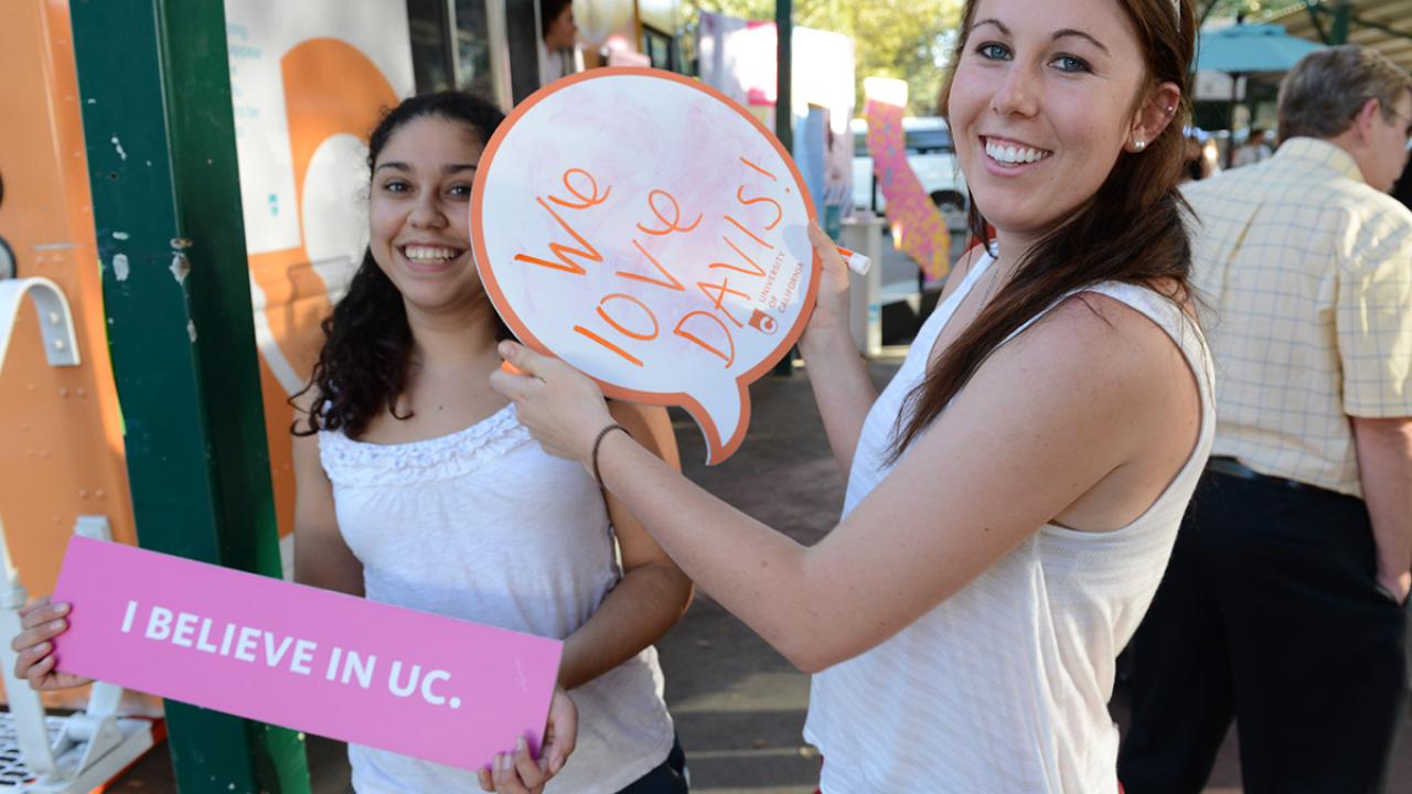 two UC Davis students pose at the Davis Farmers Market with signs saying how much they love Davis