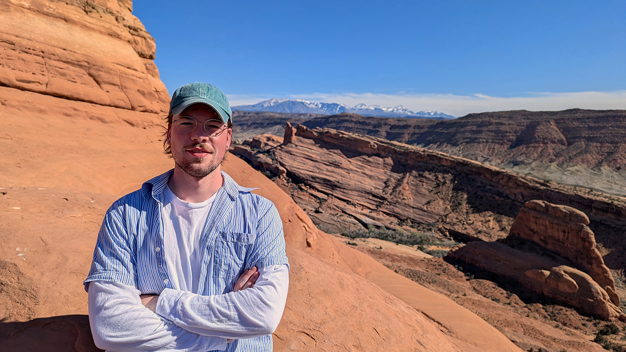 Arian,wearing a green cap, stands at a red‑rock canyon with mountains in the distance. 