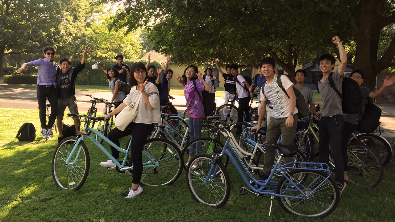 Students with bicycles on shaded grassy campus, smiling and waving