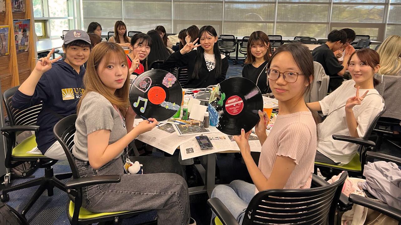 a group of international students hold up art projects that they've worked on while sitting at a table together