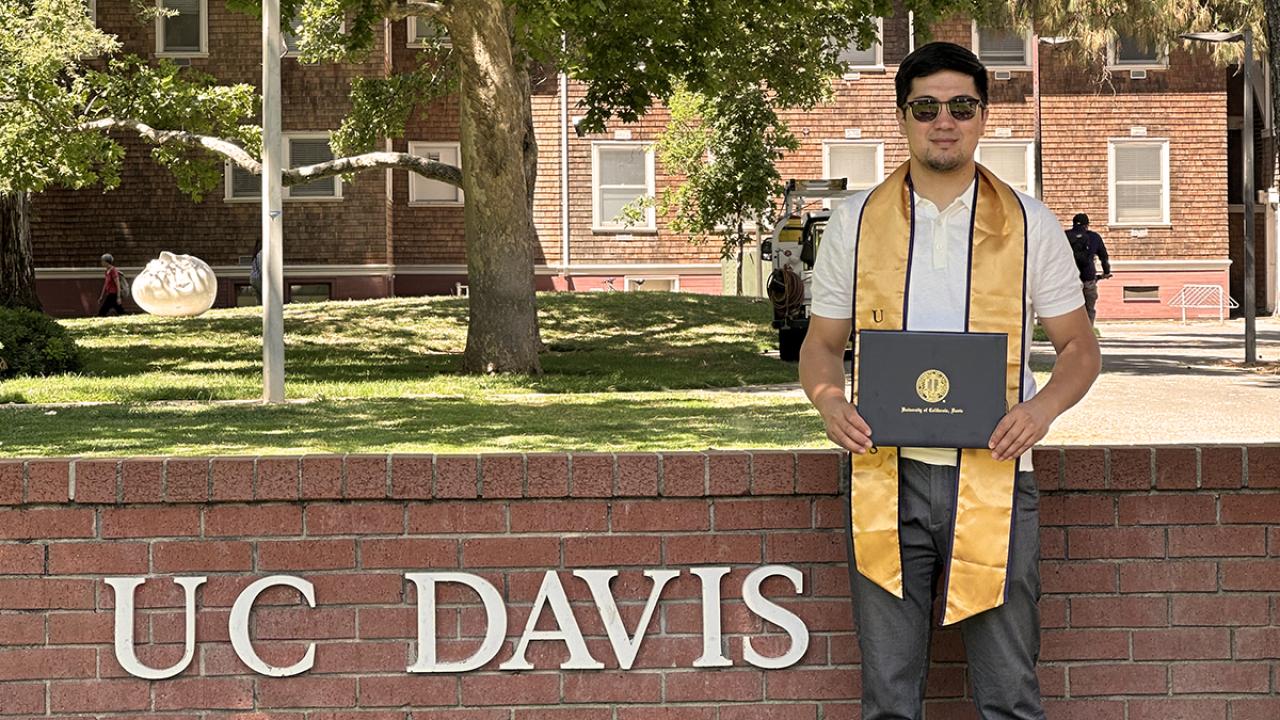 headshot of Zhankeldy Doszhanov, a Kazakh scholar who studied a UC Davis, holds his certificate of accomplishment in front of the UC Davis sign