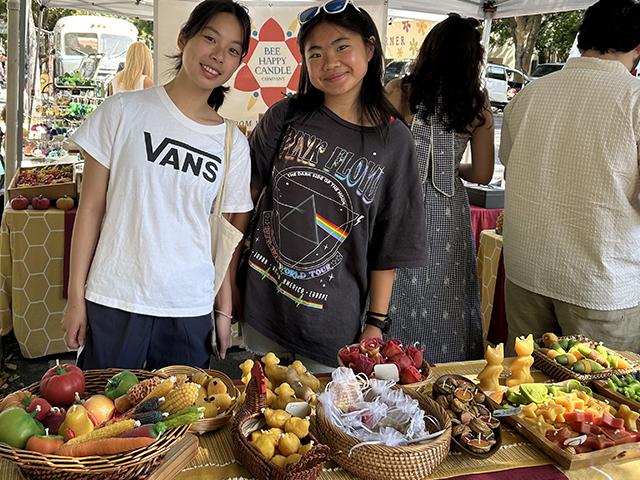 Two smiling student at an outdoor market stall with baskets of fruit and pastries.