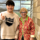 Young man giving thumbs-up beside smiling older woman in colorful cardigan, stone-tiled lobby