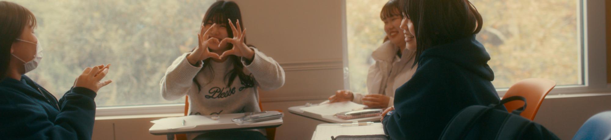 UC Davis international students sitting at desks in classroom with one person giving the hear sign with hands